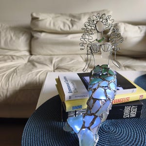 Puede incluir: Una escultura de sirena hecha de alambre y piezas de vidrio marino iridiscente. La figura presenta una cabeza y un torso formados por alambre con un top en forma de concha, y una cola construida con fragmentos de vidrio azul, verde y blanco.