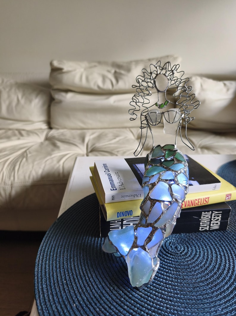 Puede incluir: Una escultura de sirena hecha de alambre y piezas de vidrio marino iridiscente. La figura presenta una cabeza y un torso formados por alambre con un top en forma de concha, y una cola construida con fragmentos de vidrio azul, verde y blanco.