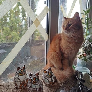 May include: Three mosaic cat figurines in shades of brown, white, and blue, sitting on a brown burlap surface. A ginger cat is sitting on the windowsill behind the figurines, looking out the window.