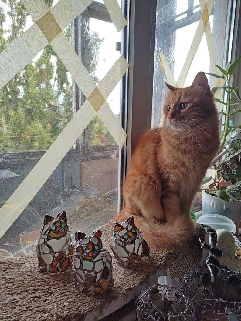 May include: Three mosaic cat figurines in shades of brown, white, and blue, sitting on a brown burlap surface. A ginger cat is sitting on the windowsill behind the figurines, looking out the window.
