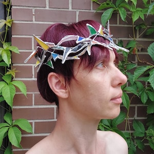 May include: A crown constructed from interconnected stained glass shards in shades of green, yellow, blue, and clear, joined by silver-toned metal solder lines. The headpiece features sharp, pointed geometric segments arranged in a circular band.