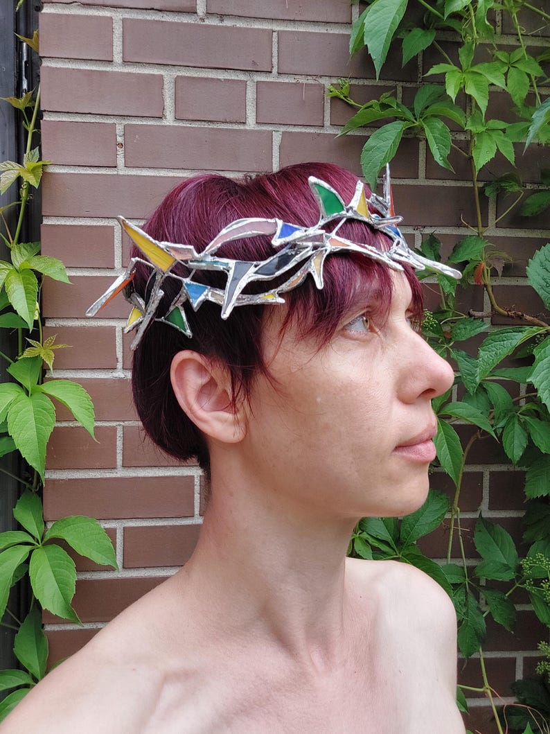 May include: A crown constructed from interconnected stained glass shards in shades of green, yellow, blue, and clear, joined by silver-toned metal solder lines. The headpiece features sharp, pointed geometric segments arranged in a circular band.