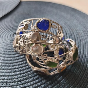 May include: A stack of silver-toned metal bracelets with colorful glass inserts. The bracelets feature blue, green, and clear glass pieces. The metalwork has a delicate, open design. The bracelets are stacked on a textured surface.