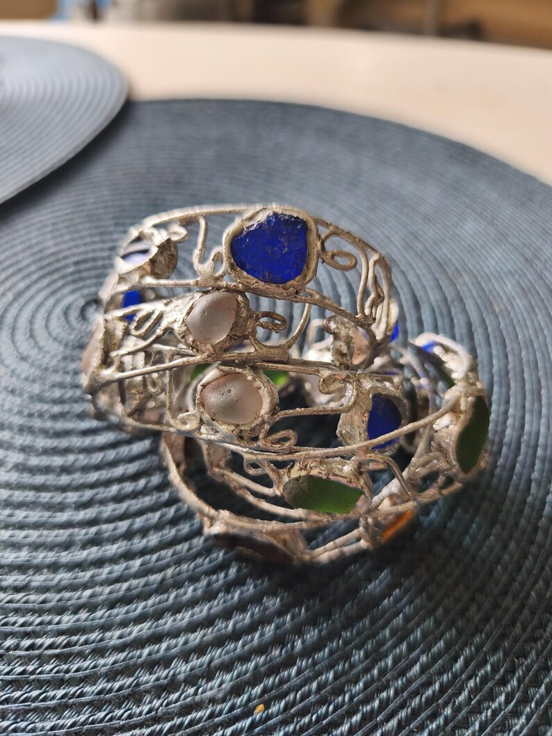 May include: A stack of silver-toned metal bracelets with colorful glass inserts. The bracelets feature blue, green, and clear glass pieces. The metalwork has a delicate, open design. The bracelets are stacked on a textured surface.