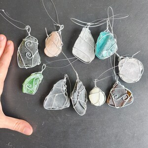 May include: A collection of sea glass pendants in various shapes and colors, including green, blue, and brown. Each piece of sea glass is wrapped in silver wire with a loop for hanging. The pendants are displayed on a dark surface.