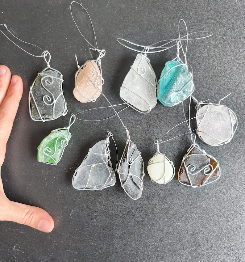 May include: A collection of sea glass pendants in various shapes and colors, including green, blue, and brown. Each piece of sea glass is wrapped in silver wire with a loop for hanging. The pendants are displayed on a dark surface.