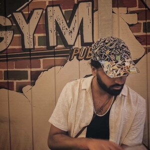 May include: A person wearing a patterned baseball cap, a cream-colored button-down shirt, and a silver chain necklace. The background features a wall with the words "GYM PUB" painted on it.