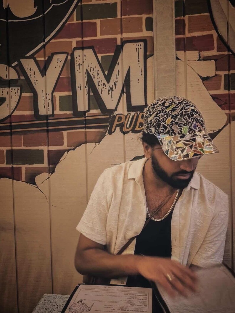 May include: A person wearing a patterned baseball cap, a cream-colored button-down shirt, and a silver chain necklace. The background features a wall with the words "GYM PUB" painted on it.