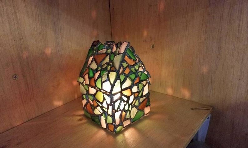 May include: A stained glass lamp with a house-shaped design. The lamp is made of small pieces of glass in shades of green, brown, and white. The lamp is lit and casting a warm glow.