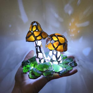 May include: A decorative mushroom lamp featuring two illuminated mushroom figures. The mushroom caps are a mosaic of yellow and brown glass, with white stems. The base is made of green glass, and the lamp emits a soft, warm glow.
