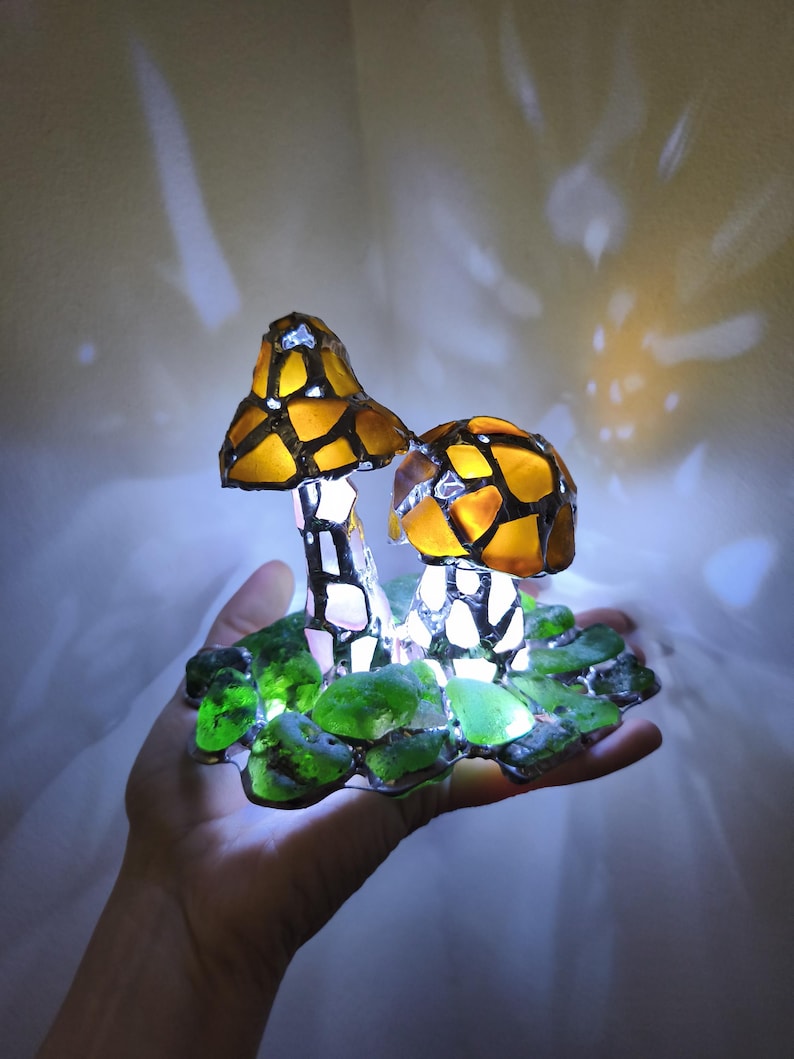 May include: A decorative mushroom lamp featuring two illuminated mushroom figures. The mushroom caps are a mosaic of yellow and brown glass, with white stems. The base is made of green glass, and the lamp emits a soft, warm glow.
