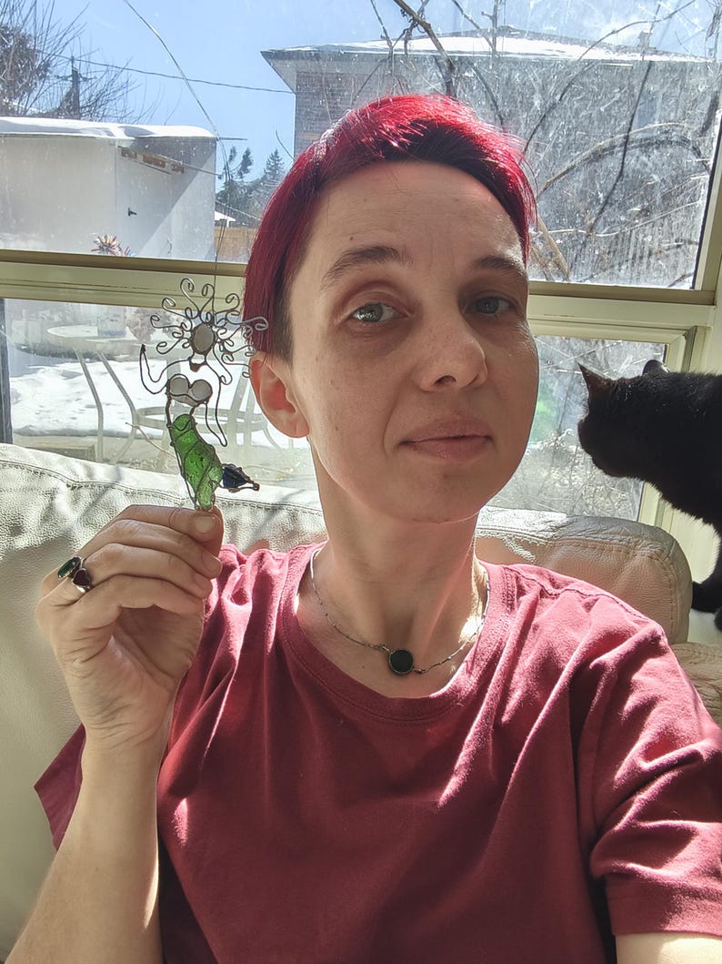 Puede incluir: Una escultura de vidrio artesanal de una figura caprichosa con un cuerpo verde y un marco de alambre plateado. La escultura se sostiene frente a una persona que lleva una camiseta roja. Un gato negro est&aacute; en el fondo.