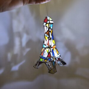 May include: A stained-glass Eiffel Tower ornament, featuring a mosaic of coloured glass pieces. The tower is suspended by a thread. The glass pieces are in shades of red, yellow, blue and green, creating a vibrant effect.