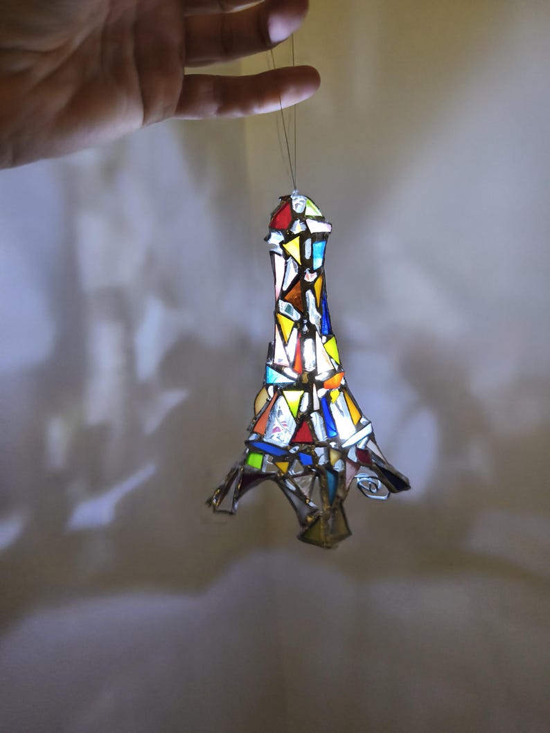 May include: A stained-glass Eiffel Tower ornament, featuring a mosaic of coloured glass pieces. The tower is suspended by a thread. The glass pieces are in shades of red, yellow, blue and green, creating a vibrant effect.