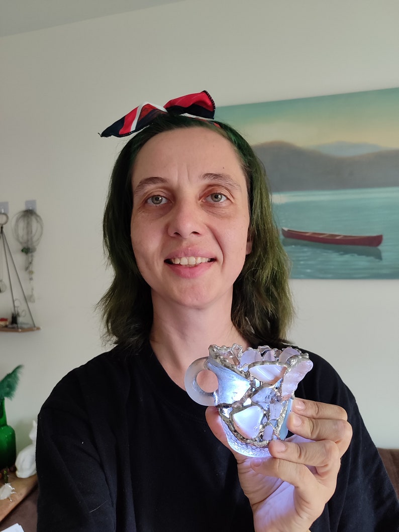 May include: A person is holding a small, clear glass sculpture that resembles a cup. The sculpture is made of many small pieces of glass that are held together with silver wire. The sculpture is lit from within, creating a warm glow.