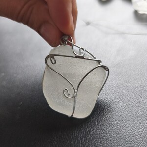 May include: A handcrafted pendant featuring a piece of frosted sea glass wrapped in silver-colored wire. The wire is shaped into decorative swirls around the edges of the sea glass, creating a unique and artistic necklace.