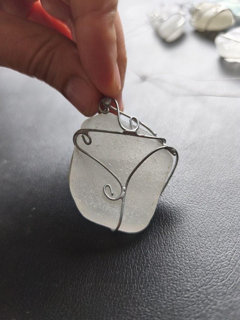May include: A handcrafted pendant featuring a piece of frosted sea glass wrapped in silver-colored wire. The wire is shaped into decorative swirls around the edges of the sea glass, creating a unique and artistic necklace.