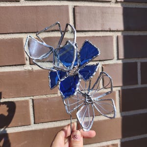 May include: A handcrafted stained glass flower sculpture with blue and clear petals, and silver wire detailing. The sculpture is held against a brick wall background, showcasing the vibrant colours and intricate design. A unique floral art piece.