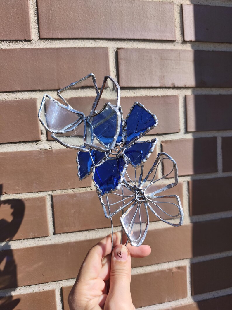 May include: A handcrafted stained glass flower sculpture with blue and clear petals, and silver wire detailing. The sculpture is held against a brick wall background, showcasing the vibrant colours and intricate design. A unique floral art piece.