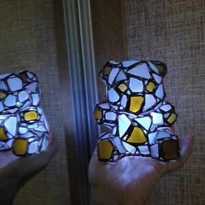 May include: A mosaic teddy bear lamp with white, yellow, and brown glass pieces. The lamp is lit from within, creating a soft glow. The bear is held in a hand, with a reflection visible in a mirror.