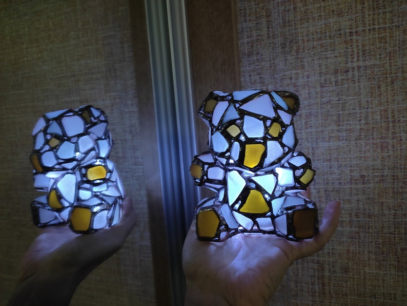 May include: A mosaic teddy bear lamp with white, yellow, and brown glass pieces. The lamp is lit from within, creating a soft glow. The bear is held in a hand, with a reflection visible in a mirror.