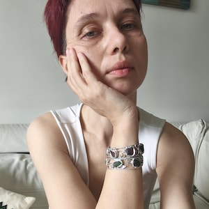 May include: Two silver bracelets with rectangular, colorful stones are stacked on a wrist. The bracelets have an open design with a decorative, swirling pattern. The stones are shades of green and red. The person is wearing a white tank top.