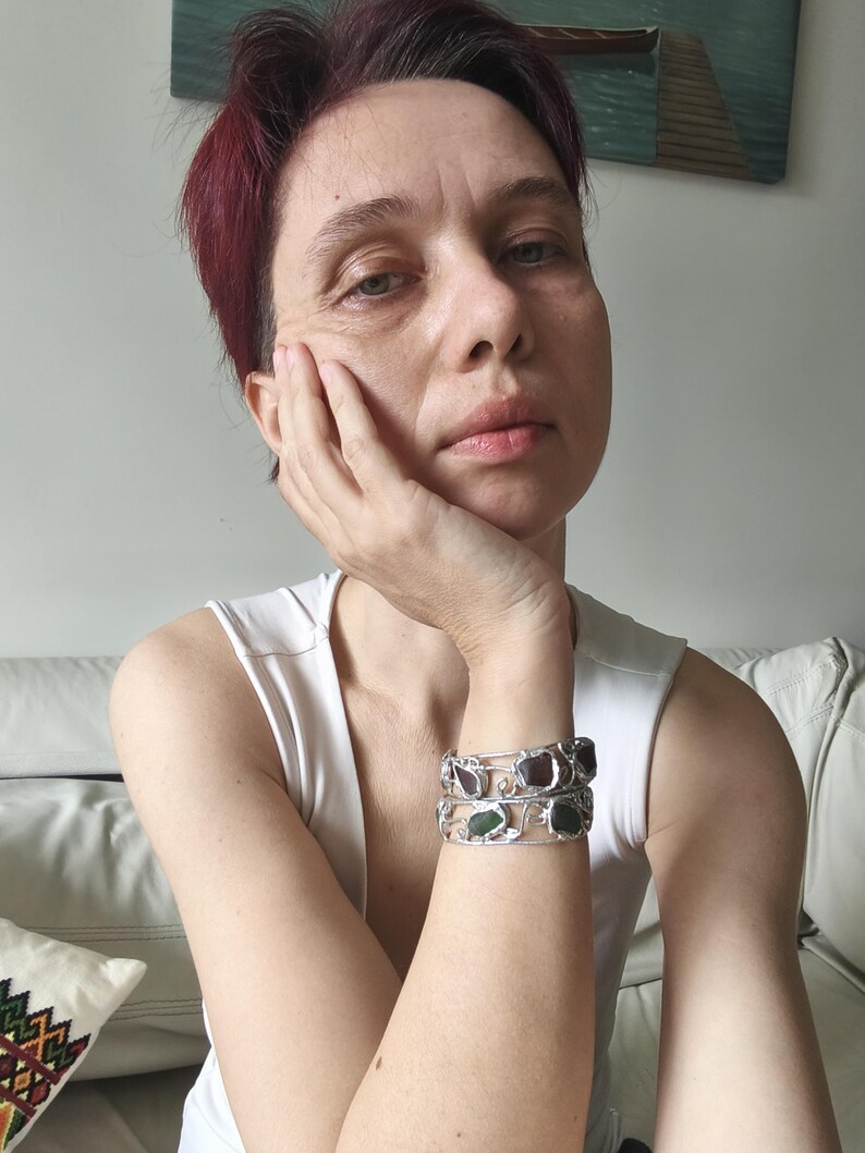 May include: Two silver bracelets with rectangular, colorful stones are stacked on a wrist. The bracelets have an open design with a decorative, swirling pattern. The stones are shades of green and red. The person is wearing a white tank top.