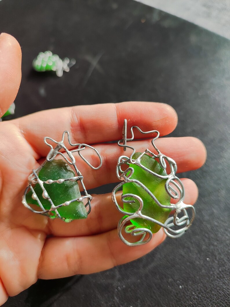 May include: Two green glass pieces are encased in silver wire, creating a unique and delicate design. The wire is twisted and shaped to resemble a tree or a plant.