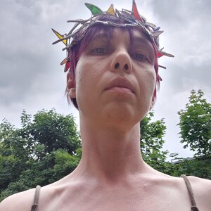 May include: A crown featuring a metallic, branch-like structure with integrated, multi-colored stained glass shards in shades of red, green, yellow, and blue. The headpiece is constructed with soldered metal joints, creating a jagged, organic silhouette.