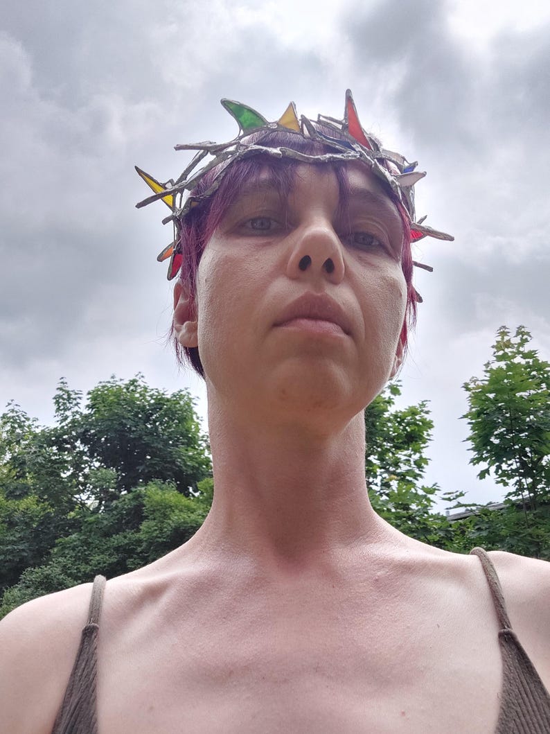 May include: A crown featuring a metallic, branch-like structure with integrated, multi-colored stained glass shards in shades of red, green, yellow, and blue. The headpiece is constructed with soldered metal joints, creating a jagged, organic silhouette.