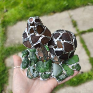 May include: A handcrafted sculpture featuring a mushroom design. The mushroom caps are made of brown and amber glass pieces, while the base is composed of green glass. The glass pieces are held together with a silver-colored material, creating a unique mosaic effect.