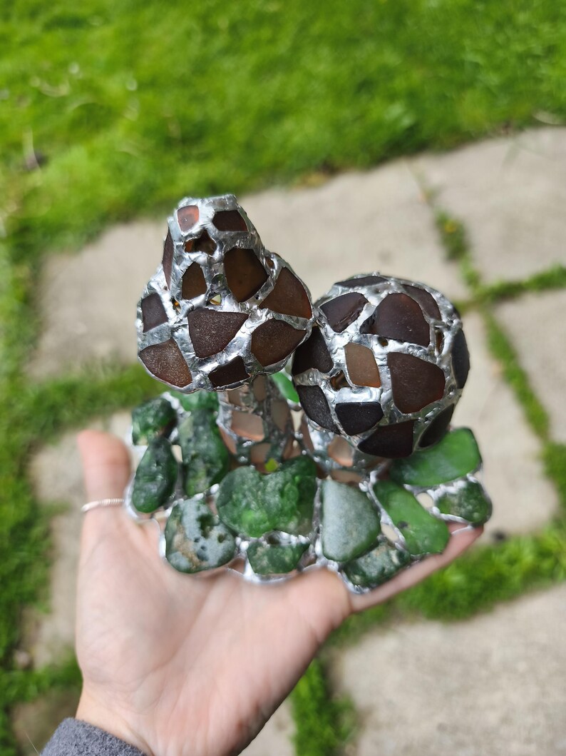 May include: A handcrafted sculpture featuring a mushroom design. The mushroom caps are made of brown and amber glass pieces, while the base is composed of green glass. The glass pieces are held together with a silver-colored material, creating a unique mosaic effect.