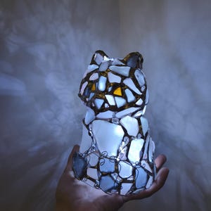 May include: A cat-shaped lamp made of white, blue, and yellow glass pieces, held in a hand. The glass is framed with a dark metal wire. The lamp is lit from within, creating a soft glow. The cat's ears are visible.