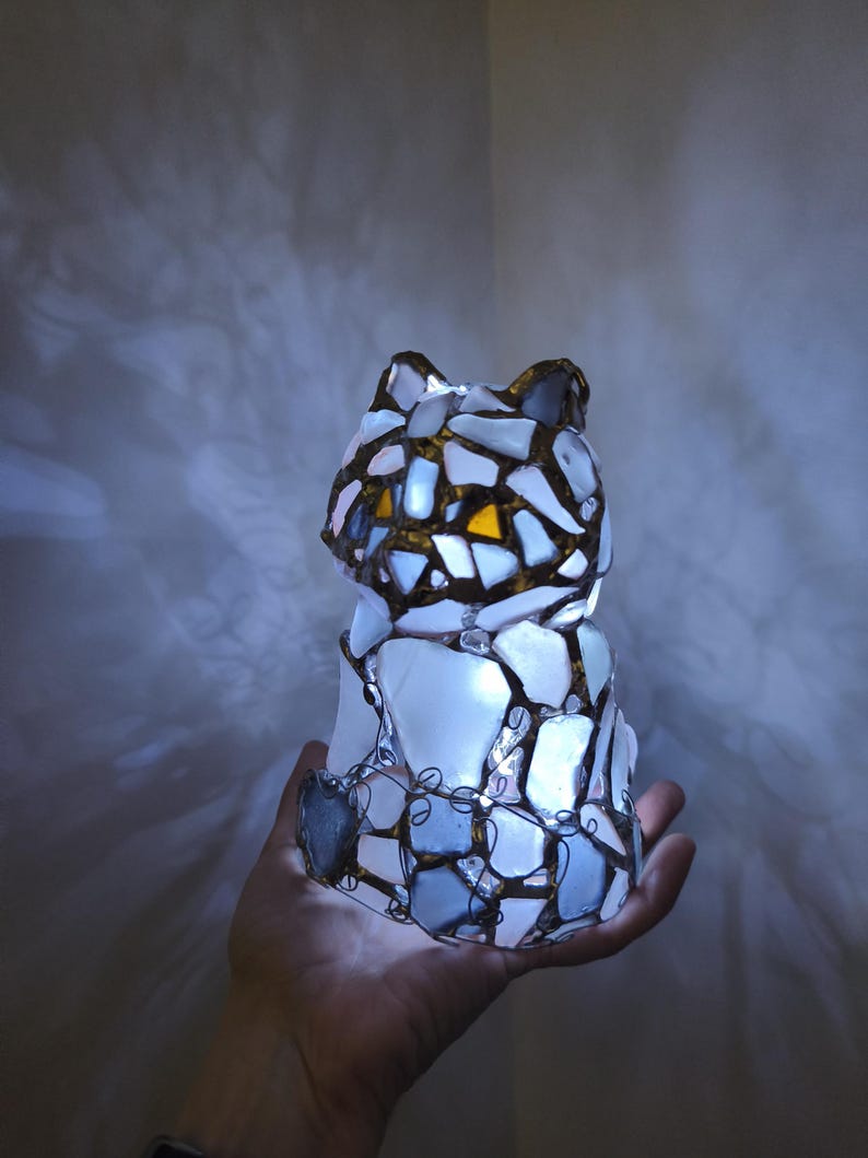 May include: A cat-shaped lamp made of white, blue, and yellow glass pieces, held in a hand. The glass is framed with a dark metal wire. The lamp is lit from within, creating a soft glow. The cat's ears are visible.