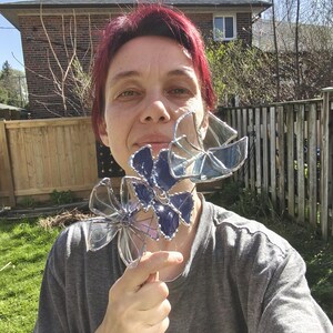 May include: A person holding two decorative glass flower sculptures. The flowers have a clear glass design with blue and purple accents. The sculptures are held in front of a green lawn and wooden fence.