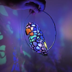 May include: A colorful, stained-glass parrot-shaped lamp with a variety of colors including orange, blue, green, and purple. The lamp is suspended by a silver metal ring. The background is a dark purple with light reflections.