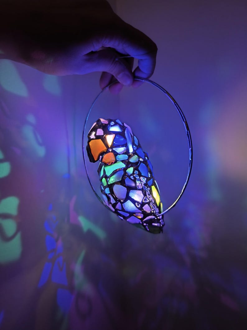 May include: A colorful, stained-glass parrot-shaped lamp with a variety of colors including orange, blue, green, and purple. The lamp is suspended by a silver metal ring. The background is a dark purple with light reflections.