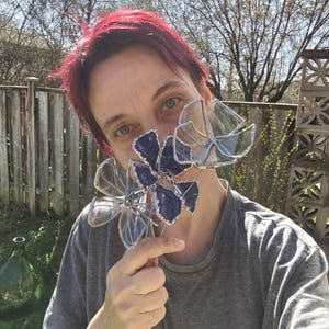 May include: Clear and blue glass flower sculptures with silver wire detailing. The flowers are held up in front of a person's face. The background includes a wooden fence, trees, and a sunny sky.