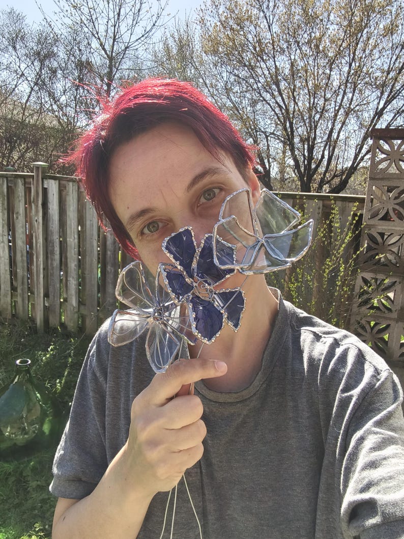 May include: Clear and blue glass flower sculptures with silver wire detailing. The flowers are held up in front of a person's face. The background includes a wooden fence, trees, and a sunny sky.