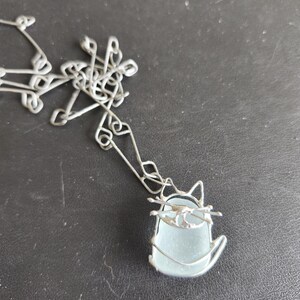 May include: A silver-toned wire cat pendant necklace with a light blue, sea glass-like body. The cat's head and whiskers are crafted from the same wire. The necklace chain is also silver-toned.