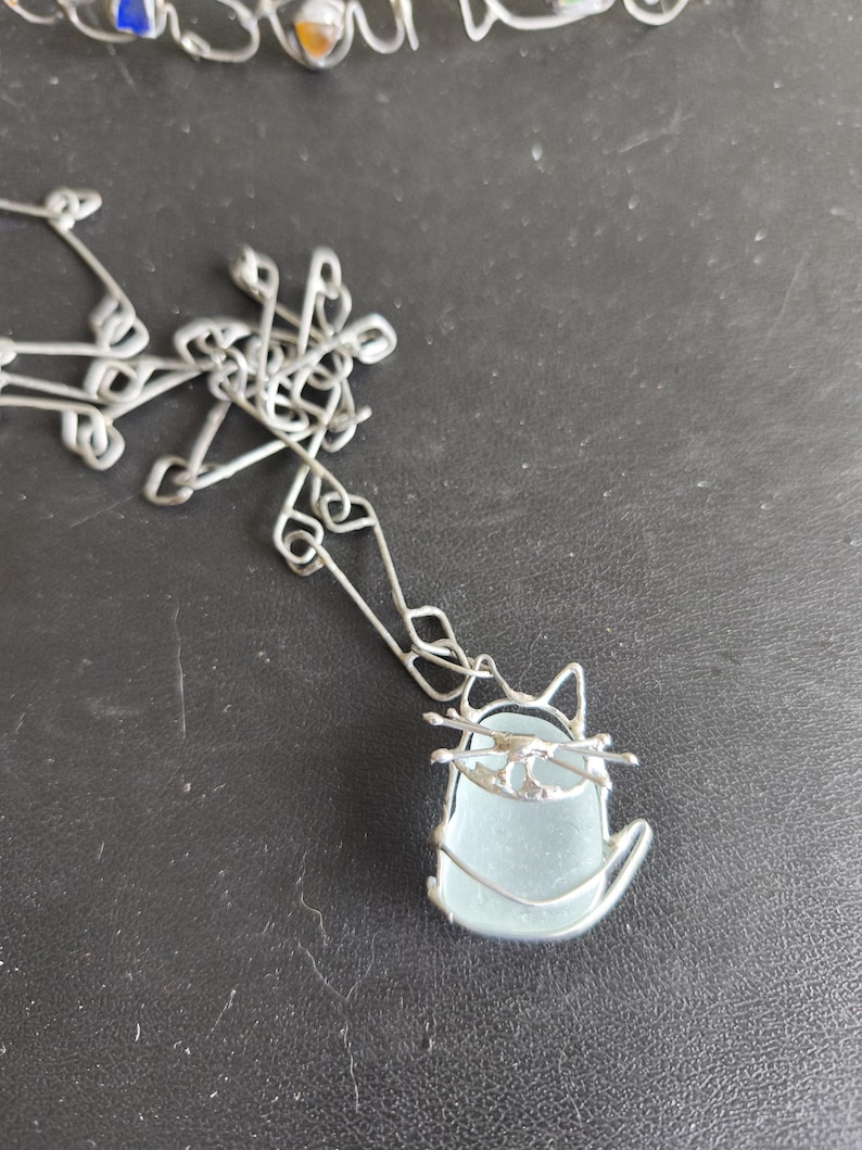 May include: A silver-toned wire cat pendant necklace with a light blue, sea glass-like body. The cat's head and whiskers are crafted from the same wire. The necklace chain is also silver-toned.
