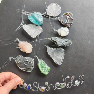May include: A collection of sea glass pendants in various colors, including blue, green, and clear, wrapped in silver wire. The word "Sea Stained Glass" is spelled out in wire with small glass pieces. The pendants are displayed on a dark surface.