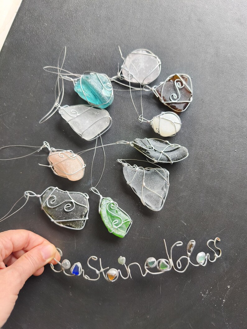 May include: A collection of sea glass pendants in various colors, including blue, green, and clear, wrapped in silver wire. The word "Sea Stained Glass" is spelled out in wire with small glass pieces. The pendants are displayed on a dark surface.