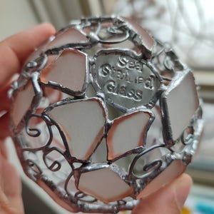 May include: A silver metal sphere made of sea glass pieces, with a label that reads "Sea Stained Glass".
