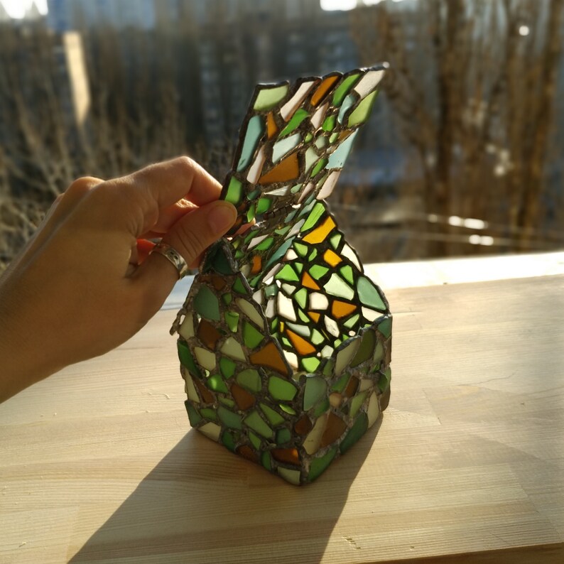 May include: A green, brown and white stained glass mosaic candle holder in the shape of a house. The top of the house is open and the holder is made of small pieces of glass.