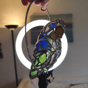 May include: A stained glass bird ornament with a metal frame. The bird features blue, green, yellow, and brown glass pieces. It is held by a metal hoop, likely for hanging. The background includes a ring light and a couch.