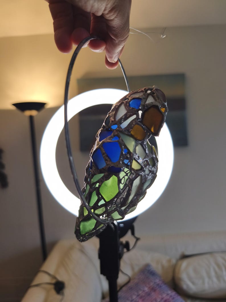 May include: A stained glass bird ornament with a metal frame. The bird features blue, green, yellow, and brown glass pieces. It is held by a metal hoop, likely for hanging. The background includes a ring light and a couch.