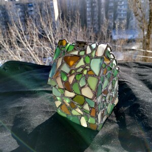 May include: A small house-shaped mosaic made from green, brown, and white glass pieces. The mosaic is set in a cement base.