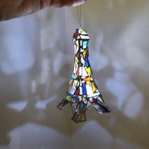 May include: A stained glass Eiffel Tower ornament in a variety of colours. The ornament is made of small pieces of glass in shades of red, blue, yellow, and white. It is suspended by a thin string.