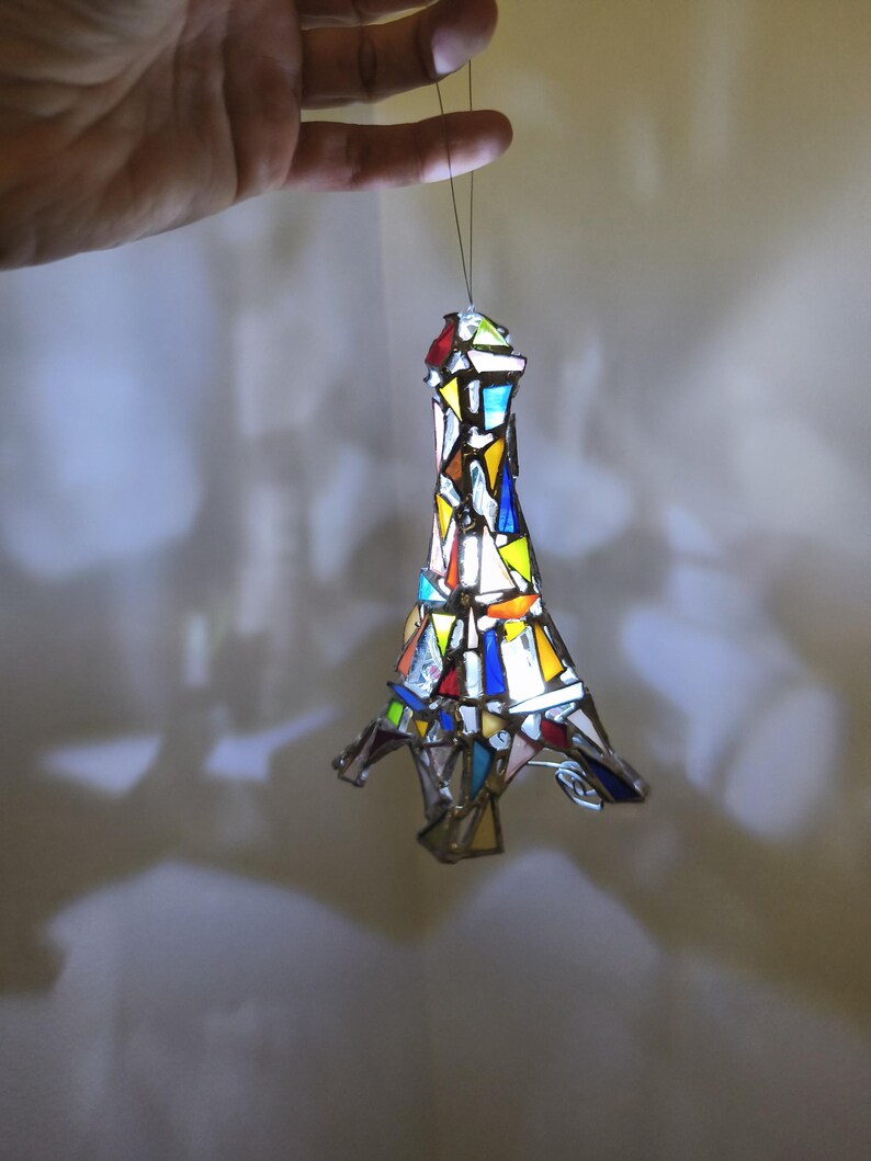 May include: A stained glass Eiffel Tower ornament in a variety of colours. The ornament is made of small pieces of glass in shades of red, blue, yellow, and white. It is suspended by a thin string.